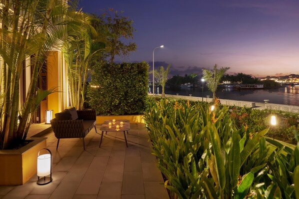 An outdoor patio area with a sofa and table, surrounded by plants and a view of a body of water at dusk. - Moire Hoi An, Vignette Collection (Hoi An)