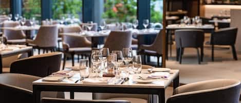 A restaurant with tables set for dining, large windows, and indoor plants.
