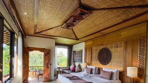 A spacious bedroom with a large bed, a wooden headboard, a bedside table with a lamp, and a view of the outdoors. - Sakti Garden Resort and Spa (Ubud)