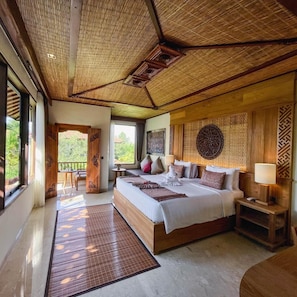 A spacious bedroom with a large bed, a wooden headboard, a bedside table with a lamp, and a view of the outdoors. - Sakti Garden Resort and Spa (Ubud)