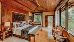 A spacious bedroom with a large bed, bedside tables, a bench, and a view of greenery through large windows. - Sakti Garden Resort and Spa (Ubud)