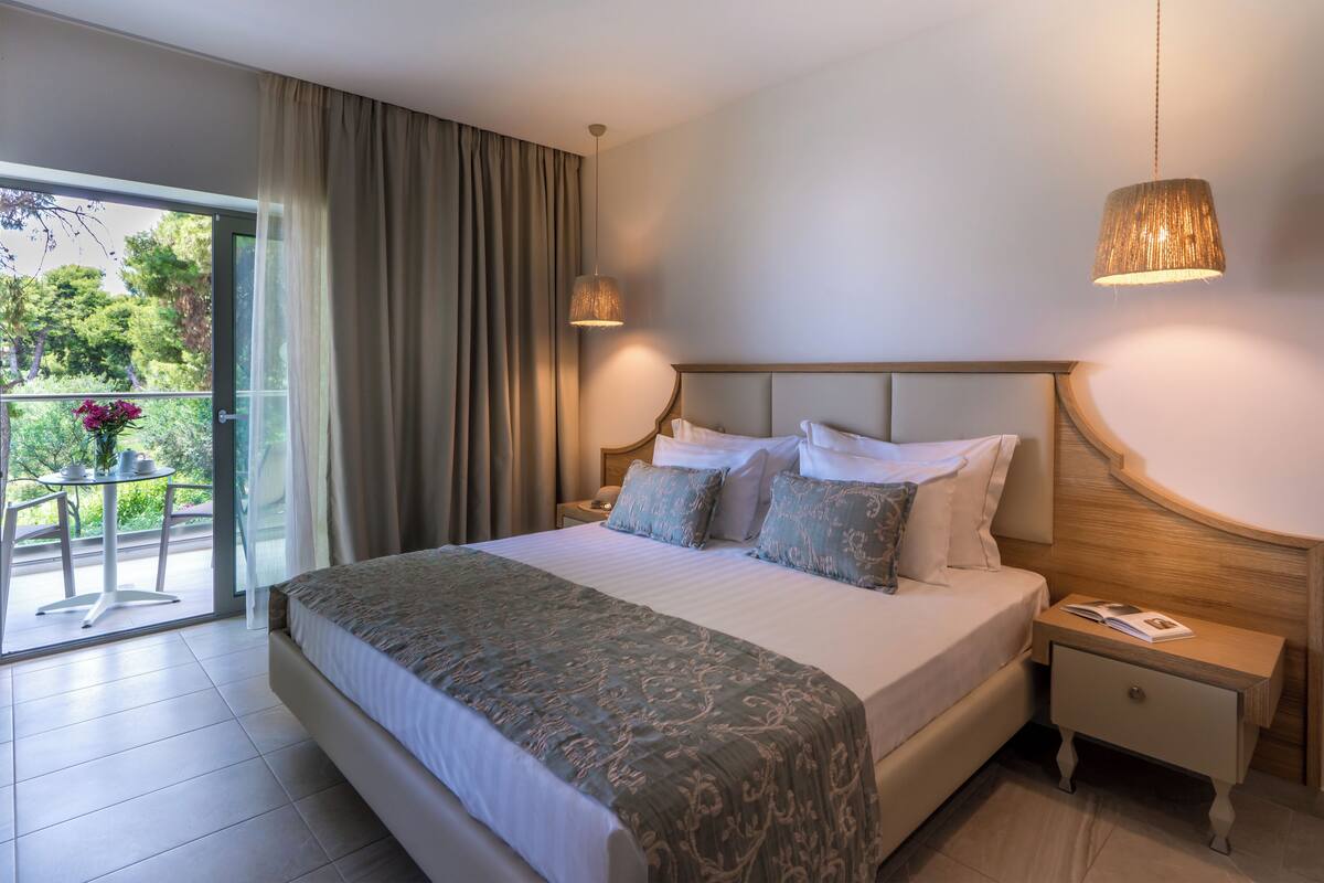 Een hotelkamer met een groot bed, nachtkastje, gordijnen voor het raam en uitzicht op groen.