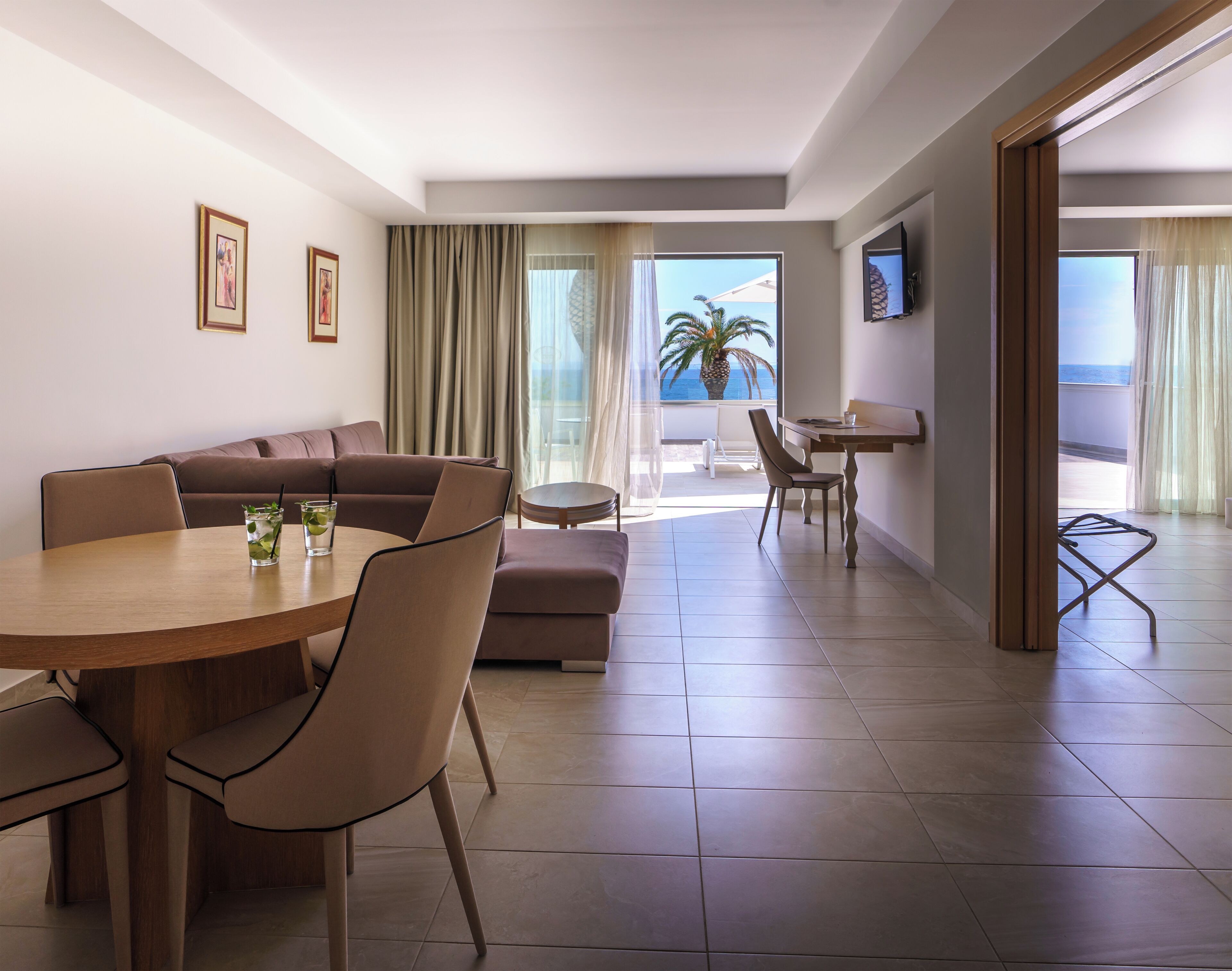 A modern hotel room with a dining area, a sofa, a small table, and a TV mounted on the wall.