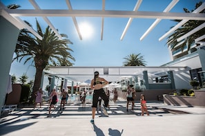 A group of people exercising outdoors under a covered area with palm trees and a pool in the background.