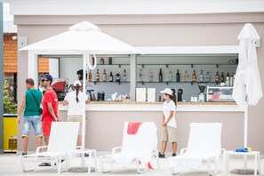 Zona de bar con sillas y sombrillas blancas, una barra con varias botellas y una persona de pie junto a la barra.