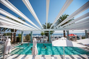 A pool area with white umbrellas and lounge chairs, a clear view of the sea, and palm trees.
