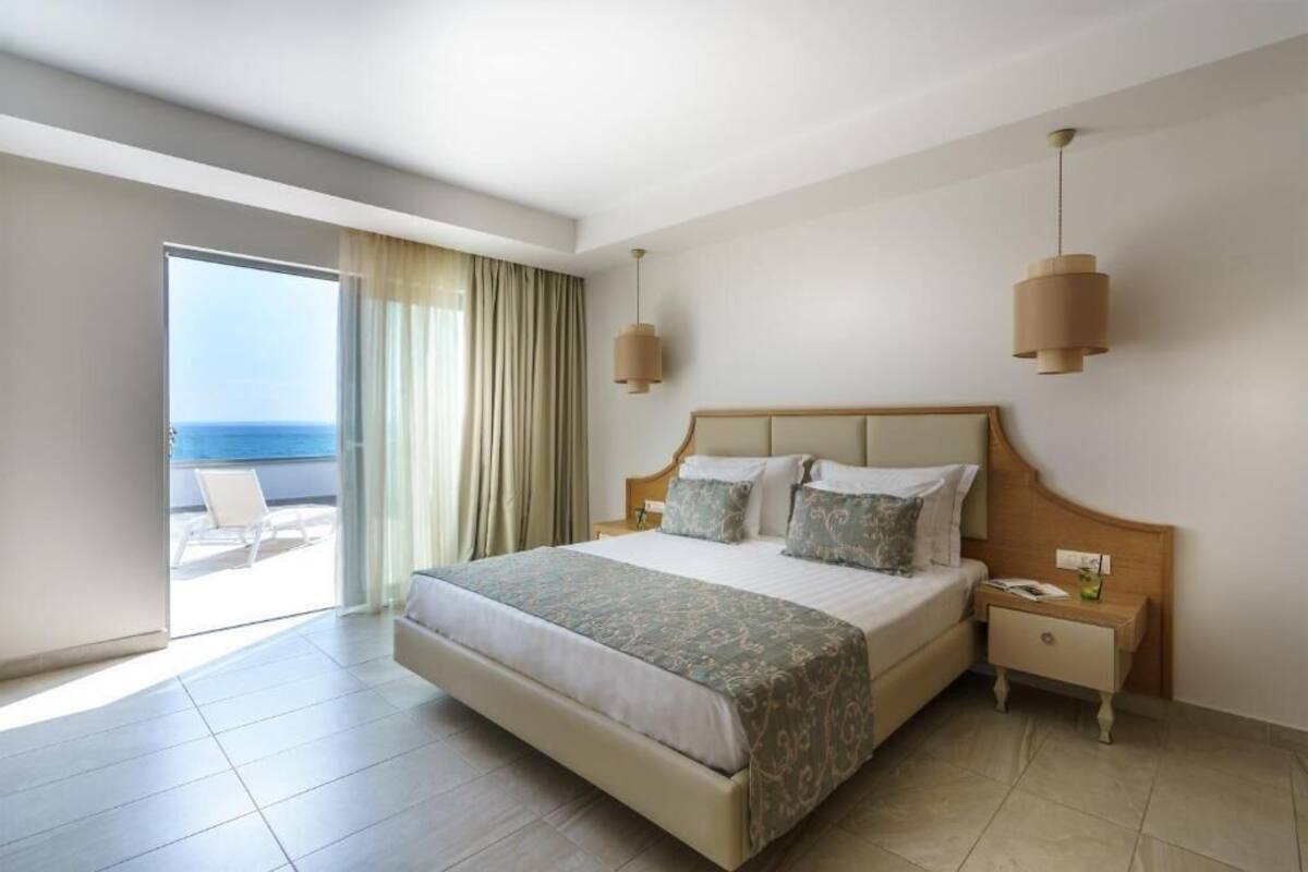 Een hotelkamer met een groot bed, een nachtkastje, een lamp, een schuifglazen deur naar een balkon en uitzicht op de zee.