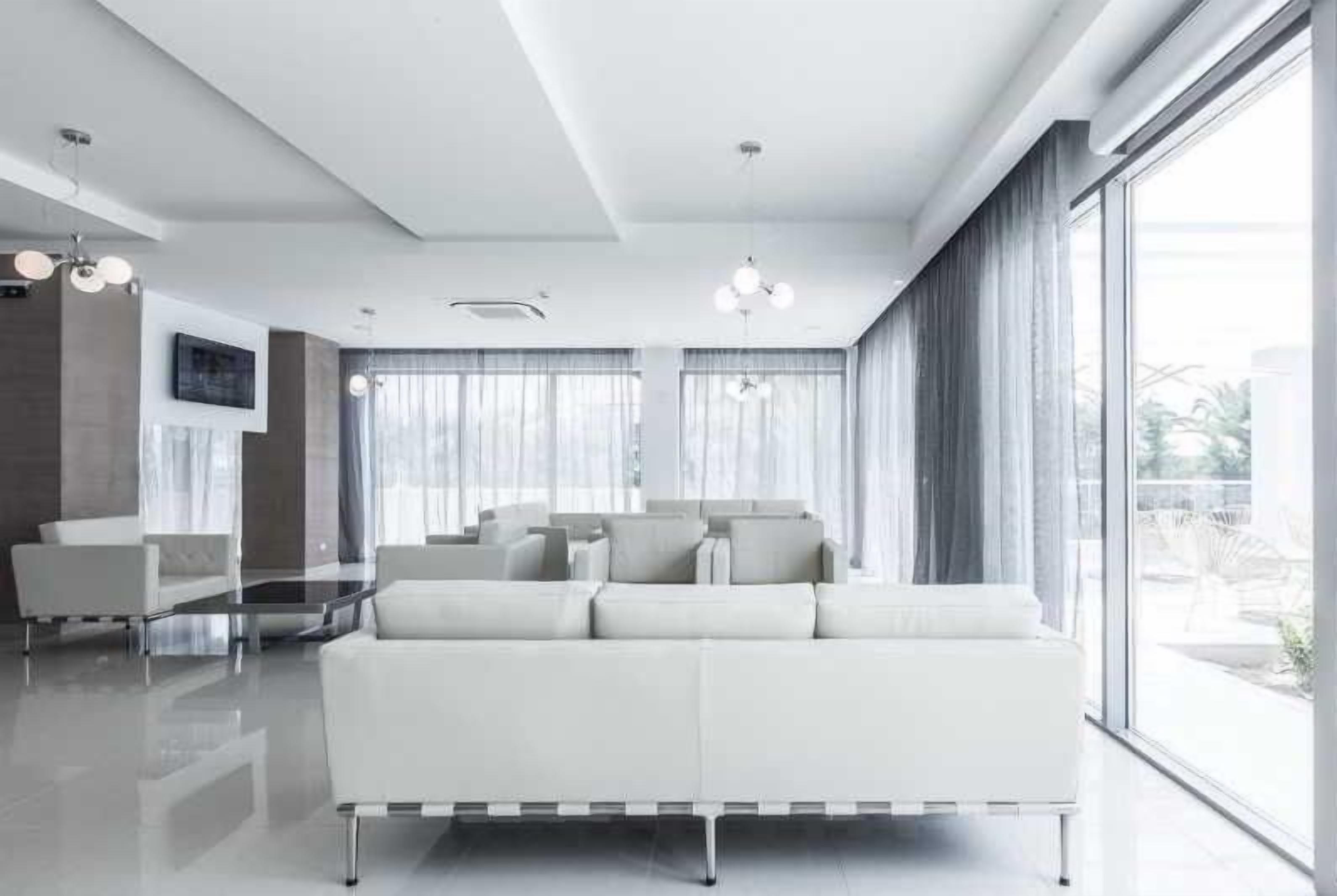 Ein modernes Wohnzimmer mit einem weißen Sofa, einem Glastisch und großen Fenstern mit Vorhängen.
