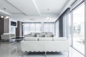 Uma sala de estar moderna com um sofá branco, uma mesa de centro de vidro e janelas grandes com cortinas.