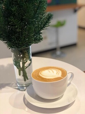 A cup of coffee with latte art on a saucer, placed on a table next to a clear glass vase with green foliage.