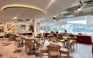 Un comedor moderno con varias mesas y sillas, un mural grande en la pared y vistas de un paisaje urbano a través de amplios ventanales.