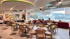 Un restaurant offrant une vue sur un bâtiment grâce à de grandes fenêtres, avec des chaises et des tables en bois, et une fresque colorée sur le mur.