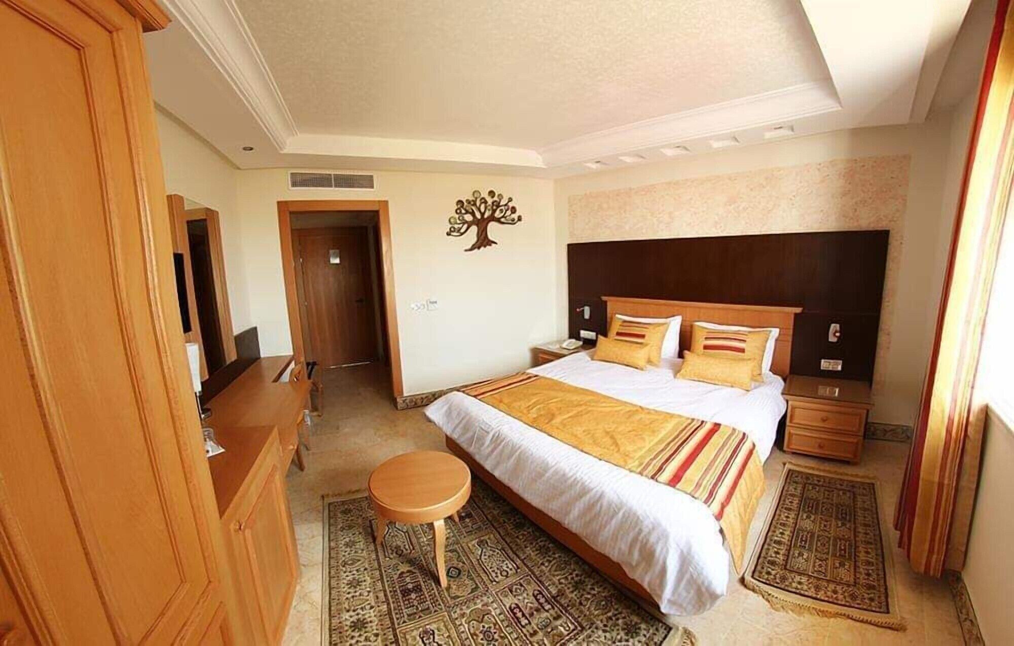 Une chambre d’hôtel comprenant un lit, un bureau, une chaise, un petit tabouret, une table de chevet, un téléphone et un élément décoratif mural.
