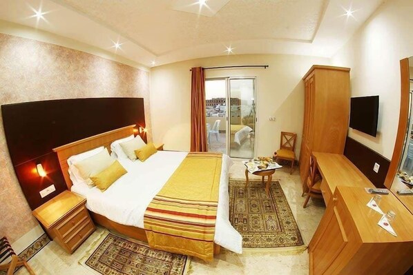 A hotel room with a large bed, a desk, a chair, a TV, and a sliding glass door to the balcony. - Julius El jem (El Djem)