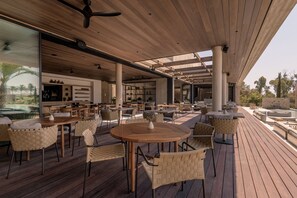 An outdoor patio with wooden tables and chairs, a ceiling fan, and a view of the surrounding landscape. - Theros All Suite Hotel (Kos)