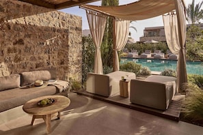 A cozy outdoor seating area with a stone wall, a pool, and a canopy. - Theros All Suite Hotel (Kos)