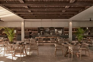 A spacious restaurant with wooden floors and furniture, a central bar area, and large windows allowing natural light. - Theros All Suite Hotel (Kos)