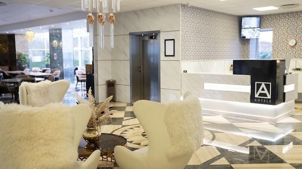 A hotel reception area with a modern design, featuring a reception desk, plush white chairs, a decorative vase, and a patterned floor. - A Hotels (Brondby)
