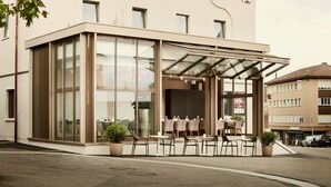 A hotel with a modern glass facade, outdoor seating area, and a clear view of the street. - Hotel Uzwil - Ihr Businesshotel (Uzwil)