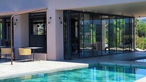 A modern outdoor pool area with a glass-enclosed patio, seating, and a clear view of the surrounding landscape. - Tetra Resort and Spa (Kefalonia)