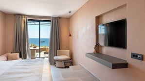 A modern hotel room with a large bed, a flat-screen TV, a small table, a chair, and a view of the sea through large windows. - Tetra Resort and Spa (Kefalonia)