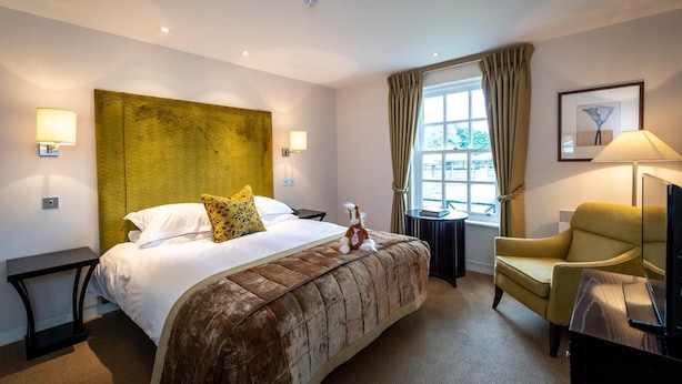Ein Hotelzimmer mit einem großen Bett, einem kleinen Tisch, einem Sessel, einem Fernseher und einem Fenster mit Vorhängen.