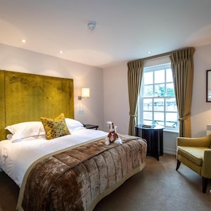 A hotel room with a large bed, a small table, a chair, a window with curtains, and a wall-mounted light fixture. - Bedford Lodge Hotel and Spa (Newmarket)