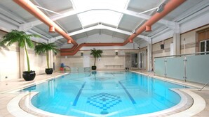 Een binnenzwembad met helderblauw water, omgeven door betegelde muren en een hoog plafond met zichtbare leidingen.