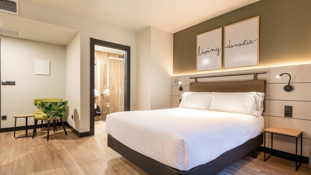 A modern hotel room with a large bed, a small desk, and a chair. There are two framed signs on the wall and a door leading to another room.