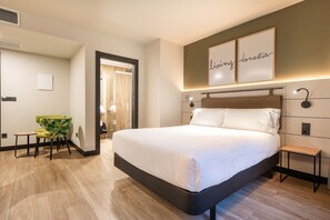 A modern hotel room with a large bed, a small desk, and a chair. There are two framed signs on the wall and a door leading to another room. - Hotel Bed4u Zurriola (San Sebastián)