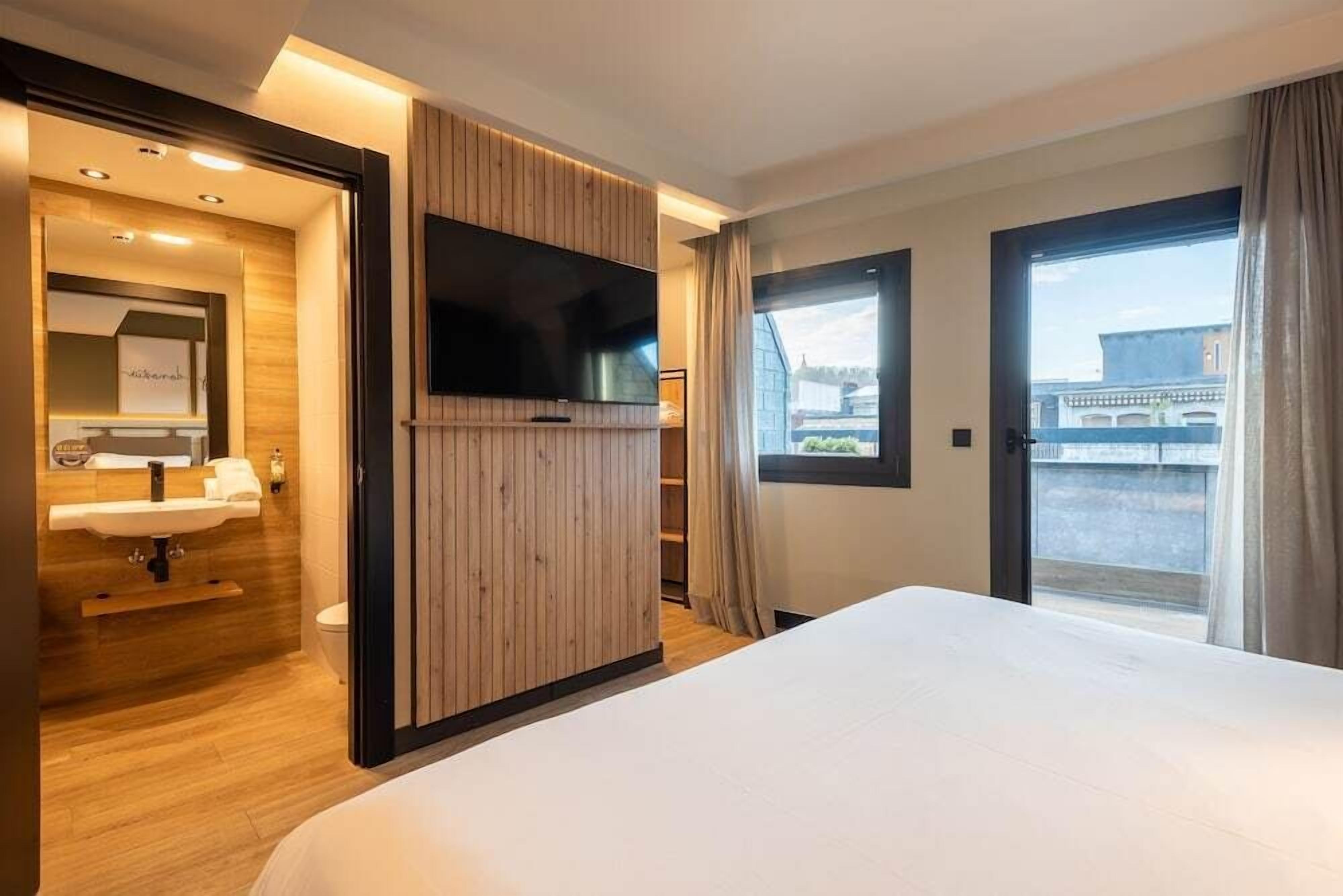 Una habitación de hotel moderna con una cama grande, un televisor de pantalla plana, un panel de pared de madera, un baño con lavabo y espejo, y un balcón con vistas.
