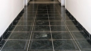 Un pasillo largo y estrecho con paredes blancas y pisos de baldosas negras. Hay ventanas a ambos lados del pasillo, y se observa una lámpara en el techo.