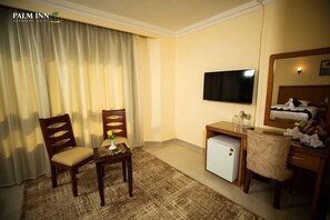 A hotel room with a bed, a TV, a desk with a computer, a chair, a small table with a vase, and a window with curtains. - Palm Inn Hotel (Hurghada)