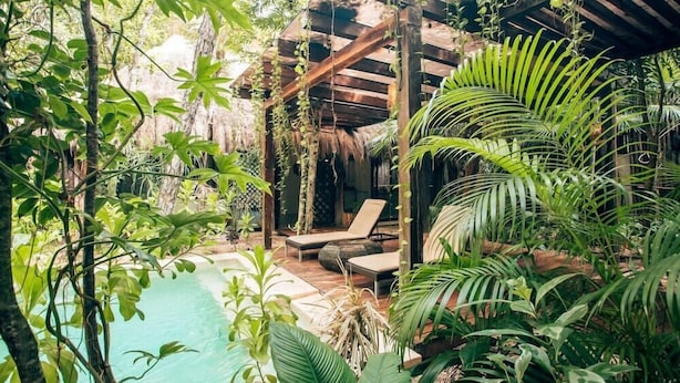 Zona junto a la piscina con sillones de descanso, una pérgola de madera y exuberante vegetación tropical.