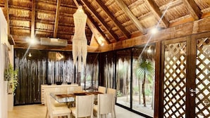 Un comedor con una mesa redonda y sillas, techo de paja y vista a palmeras a través de una puerta con celosías.