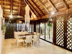 Un comedor con una mesa redonda y sillas, techo de paja y vista a palmeras a través de una puerta con celosías.