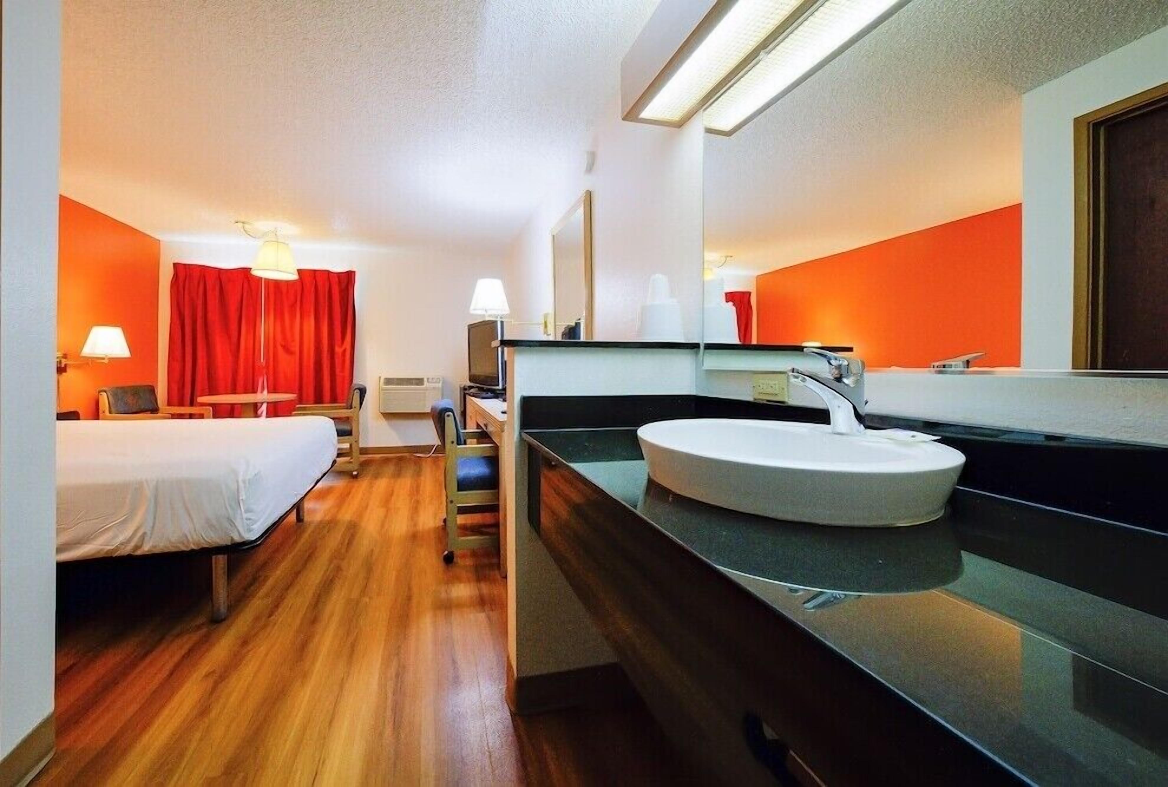 Una habitación de hotel con cama, escritorio, silla, un espejo grande y un lavamanos redondo.