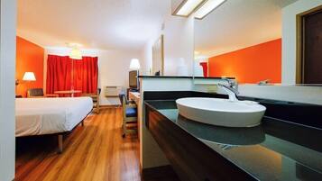 Una habitación de hotel con cama, escritorio, silla, un espejo grande y un lavamanos redondo.