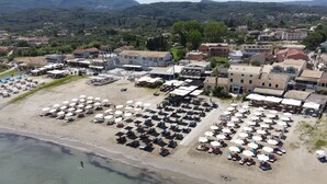 An aerial view of a beach with rows of sunbeds and umbrellas, a building with a flat roof, and a mountainous backdrop. - Aqua Luxury Suites (Corfu)