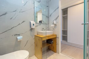 A bathroom with a marble wall, a wooden vanity, a white sink, a mirror, a glass shower enclosure, and a toilet. - Aqua Luxury Suites (Corfu)