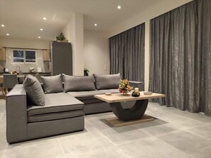A modern living room with a grey sectional sofa, a wooden coffee table, and a kitchen in the background. - Aqua Luxury Suites (Corfu)