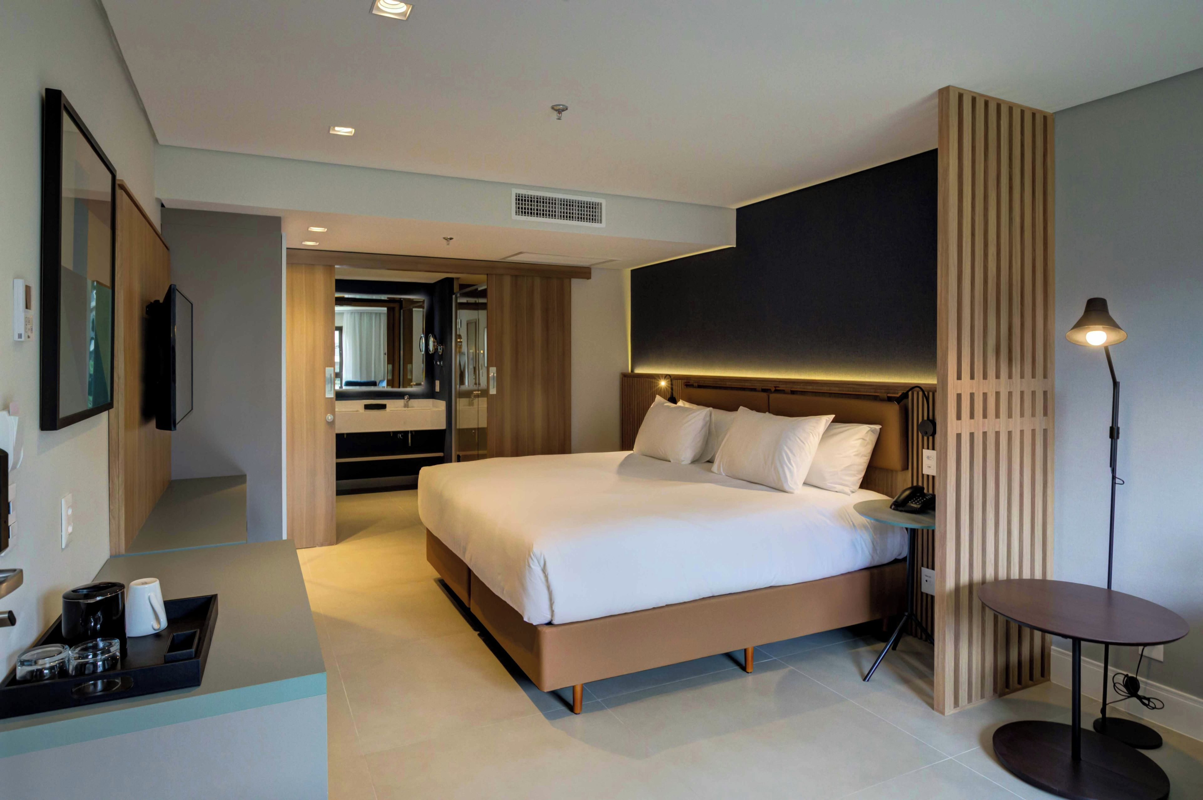 A modern hotel room with a large bed, bedside table, and a small round table with a lamp.