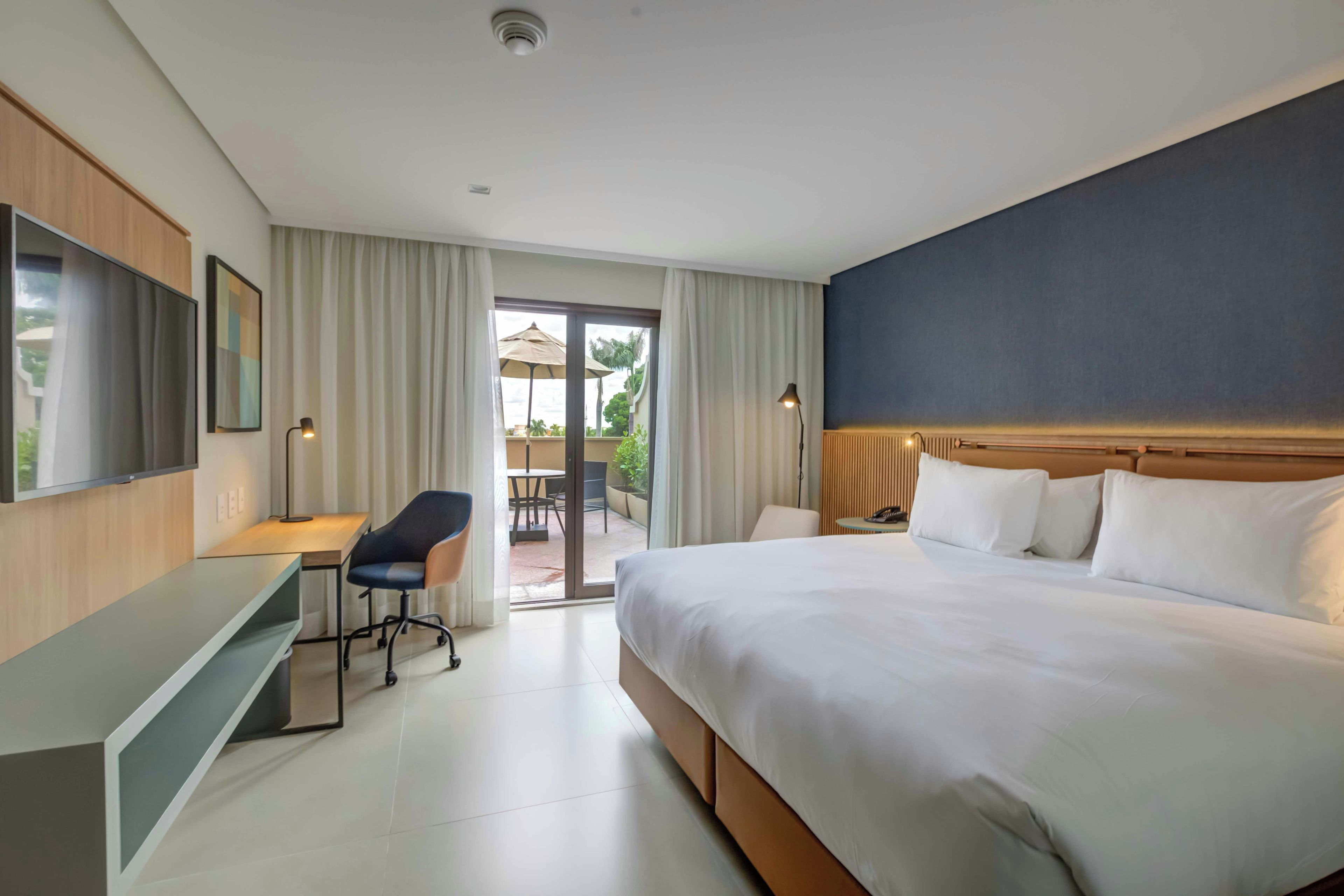 A hotel room with a large bed, a desk, a chair, a TV, and a view of the outdoors.