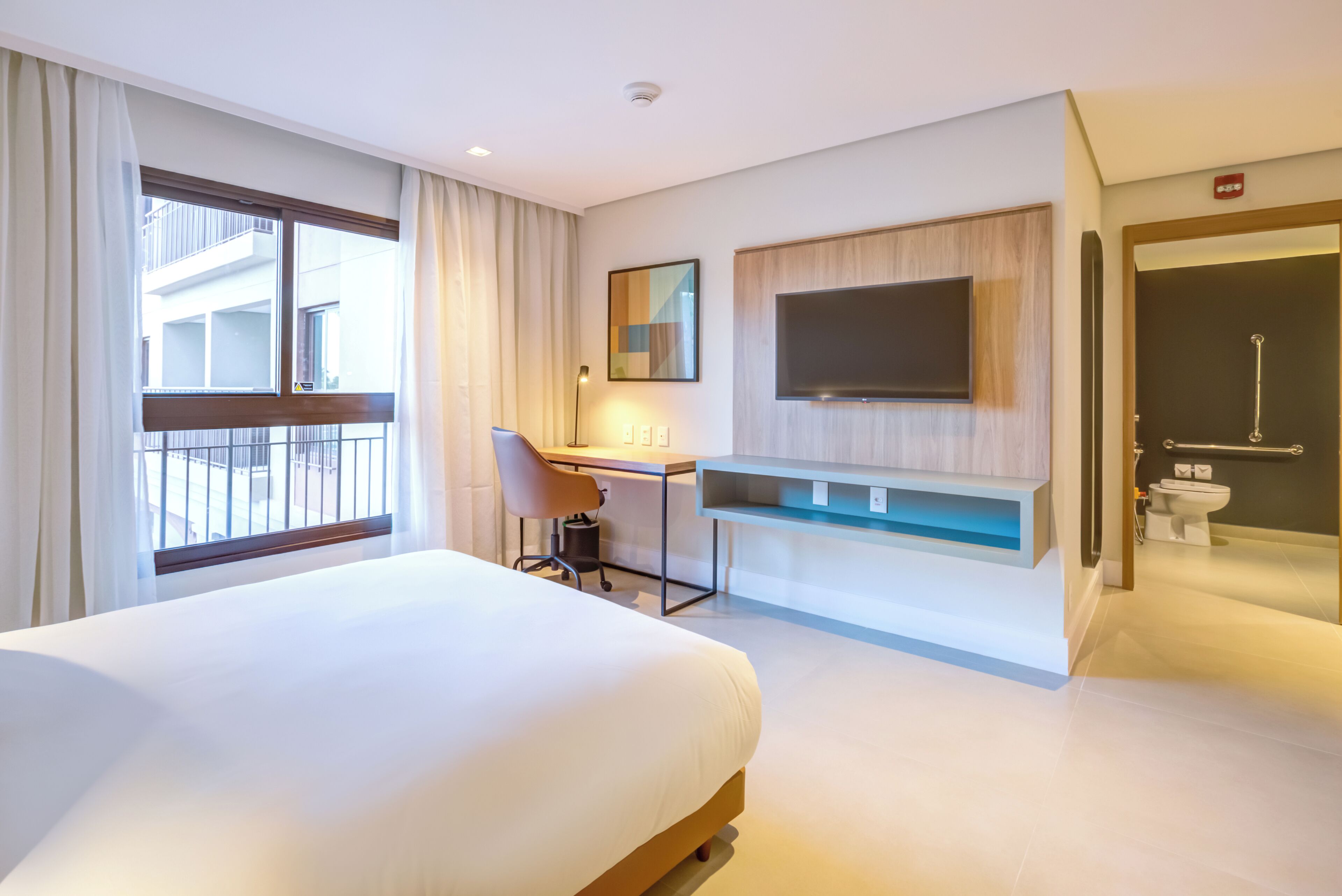 Quarto de hotel com cama, escrivaninha, televisão e banheiro.