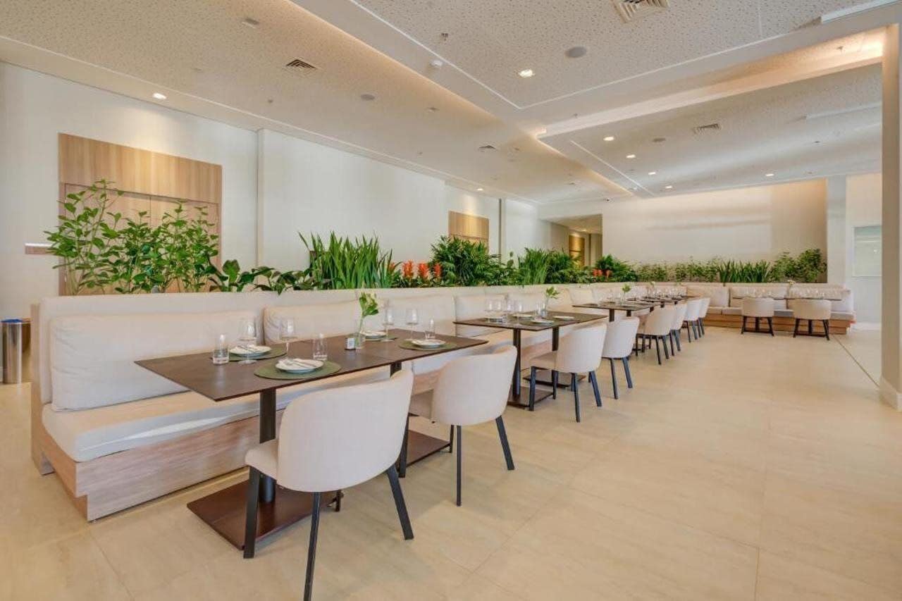 Uma área de refeições moderna, com mesas compridas e cadeiras brancas, cercada por plantas verdes e um sofá branco.