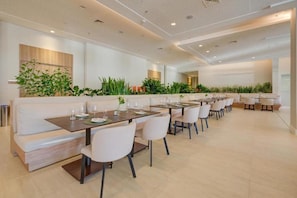 A modern dining area with long tables and white chairs, surrounded by green plants and a white sofa. - Hilton Garden Inn Sao Jose do Rio Preto (São José do Rio Preto)