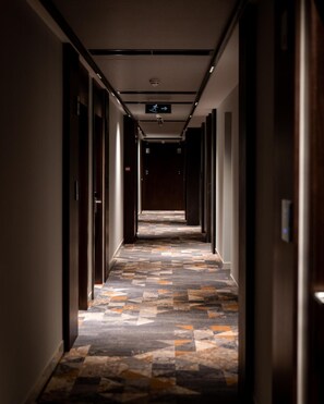 A hotel hallway with a patterned floor, doors on both sides, and a directional sign on the ceiling. - Emery Hotel Pristina (Pristina)
