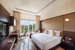 A hotel room with a large bed, a desk with a TV, a chair, and a view of greenery through the window. - Accord Chrome Airport Hotel (Chennai)