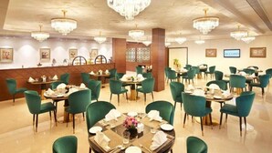 A hotel dining area with multiple round tables, green upholstered chairs, and chandeliers. - Accord Chrome Airport Hotel (Chennai)