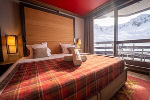 A hotel room with a large bed, two bedside lamps, a view of mountains, and a balcony with chairs.